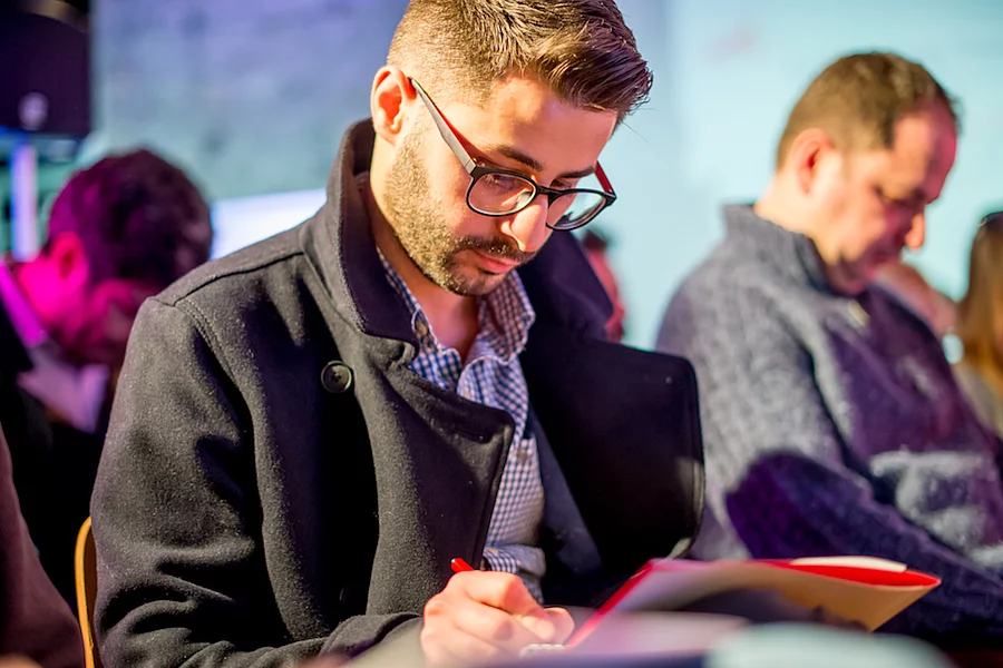 This image shows a person focused on writing in a notebook while wearing a dark pea coat and glasses.