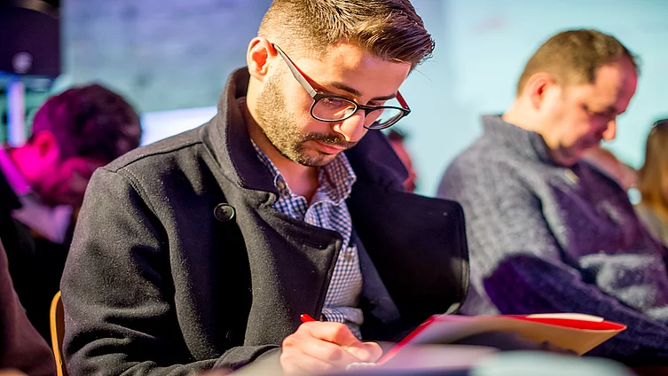 This image shows a person focused on writing in a notebook while wearing a dark pea coat and glasses.